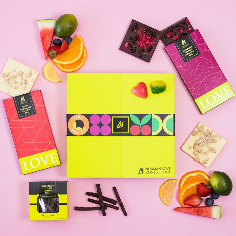 Truffle box with fruit flavored candies on pink background. Fruit, square pieces of dark chocolate raspberry inclusion bar, white chocolate passionfruit inclusion bar, and chocolate covered candied orange peels surround brightly colored truffle box. 